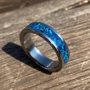 Op de afbeelding: Een zilveren ring met een blauwe opaal inlay. De ring is een eenvoudige band met een dunne lijn van blauwe opaal stenen die in het metaal zijn gezet.