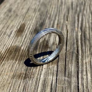 May include: A silver ring with a textured, patterned surface. The ring is made of metal and has a smooth, polished finish.