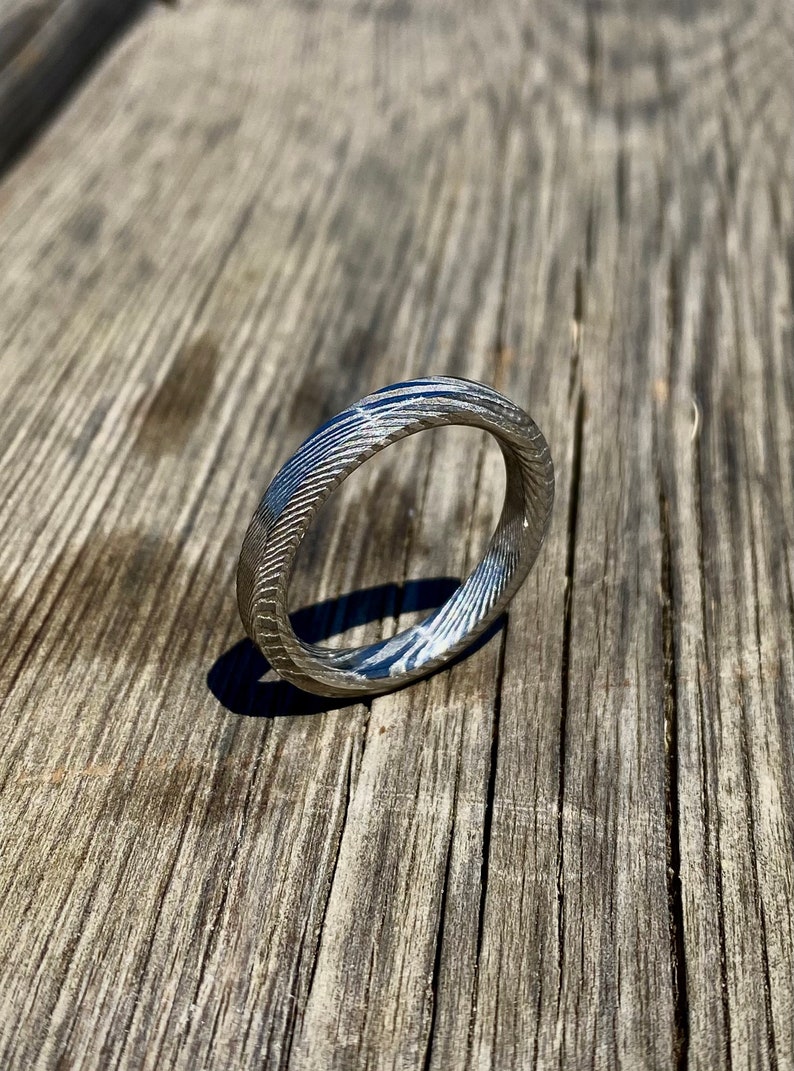 May include: A silver ring with a textured, patterned surface. The ring is made of metal and has a smooth, polished finish.