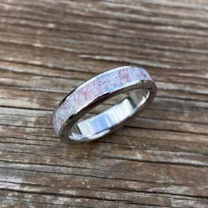 以下が含まれることがあります： ピンクと白の斑点模様の素材を使用したシルバーリング。リングは木製の背景に配置され、磨かれた金属と象嵌デザインのユニークな質感を際立たせています。リングのデザインはシンプルでエレガントです。