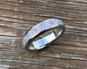 Puede incluir: Un anillo plateado con una banda de material moteado rosa y blanco. El anillo se encuentra sobre un fondo de madera, mostrando su metal pulido y la textura única del diseño incrustado. El diseño del anillo es simple y elegante.