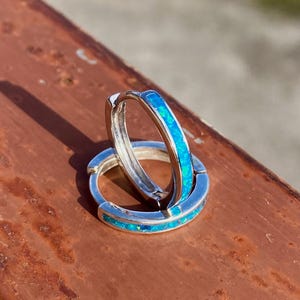Könnte beinhalten: Zwei silberne Creolen mit leuchtend blauem Opal-Inlay. Die Ohrringe werden auf einer verwitterten, rotbraunen Oberfläche präsentiert, die das Sonnenlicht einfängt. Die Farbe des Opals kontrastiert mit dem Silber und erzeugt einen auffälligen Effekt.