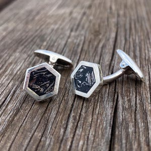 Puede incluir: Un par de gemelos plateados con diseño hexagonal. Cada gemelo presenta un centro negro con un patrón texturizado. Los gemelos se muestran sobre una superficie de madera.