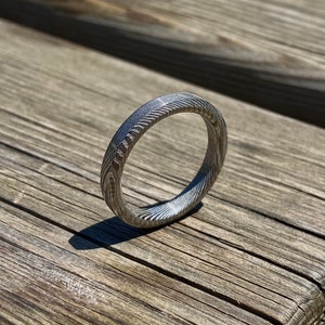 May include: A silver ring with a textured, patterned surface. The ring is made of metal and has a simple, modern design.