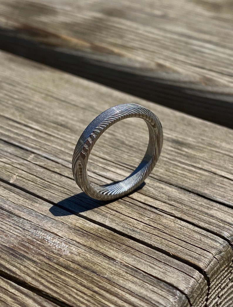 May include: A silver ring with a textured, patterned surface. The ring is made of metal and has a simple, modern design.