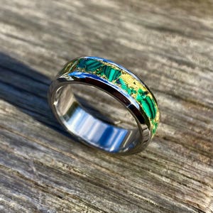 Puede incluir: Un anillo de plata con una incrustación verde y dorada. La incrustación está hecha de malaquita y escamas de oro.