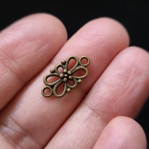 Puede incluir: Un pequeño conector de color bronce antiguo con un diseño floral ornamentado. El conector tiene dos bucles en cada extremo y un patrón central en forma de flor. La pieza se sostiene en una mano.