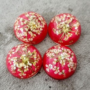 Puede incluir: Cuatro cabujones redondos de resina con fondo rojo y flores blancas prensadas en su interior. Las flores están dispuestas en forma circular.