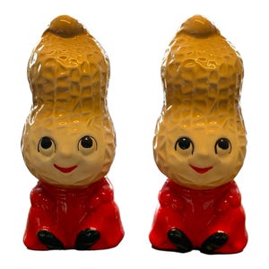 Puede incluir: Un par de saleros y pimenteros de cerámica con forma de cacahuete. Los saleros tienen forma de cacahuete con ropa roja y caras sonrientes.