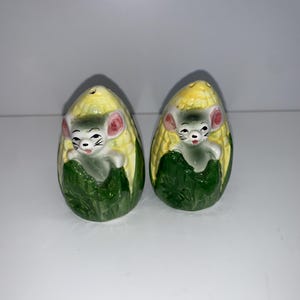 May include: A set of two ceramic salt and pepper shakers shaped like ears of corn with a cute mouse peeking out of each ear. The corn is yellow and green, and the mouse is gray with pink ears.