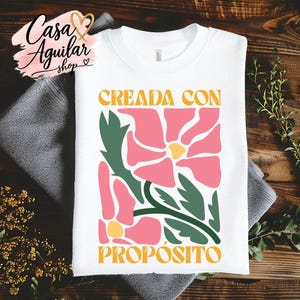 May include: A white crew neck t-shirt featuring a graphic print of pink flowers with yellow centers and dark green leaves. The text "CREADA CON" appears in yellow above the floral design, and the word "PROPÓSITO" is printed in yellow below it.