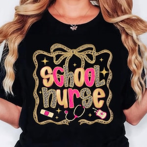 May include: Black t-shirt with a gold and pink graphic that says "school nurse." The text has a leopard print pattern. The design includes a bow, stars, a bandage, a stethoscope, and a bottle of hand sanitizer.