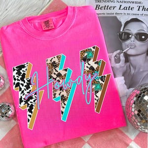 May include: A bright pink t-shirt with the word "Howdy" in a turquoise script. The word is overlaid on three-dimensional lightning bolt graphics with cow print, leopard print, and turquoise accents. The shirt is a Comfort Colors brand.