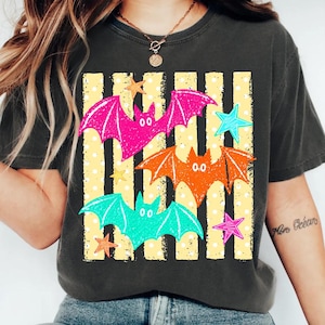 Op de afbeelding: Een antracietgrijs T-shirt met korte mouwen en een grafische print van drie gestileerde vleermuizen in felroze, oranje en turquoise. Het ontwerp bevat een achtergrond van verticale gele strepen met witte stippen en diverse gekleurde sterren.