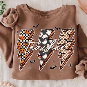May include: Brown sweatshirt with a Halloween-themed design. The design features three lightning bolt shapes with the word "Teacher" in the center. The lightning bolts have jack-o'-lanterns, ghosts, and bats. Additional bats and stars are scattered around the design.