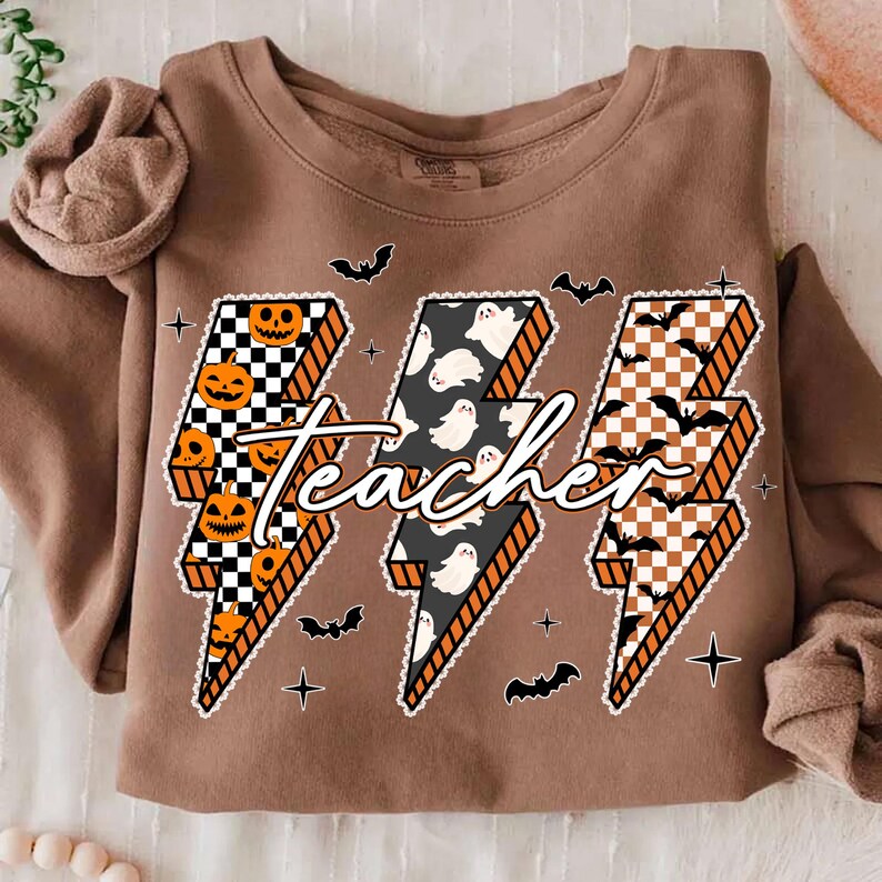 May include: Brown sweatshirt with a Halloween-themed design. The design features three lightning bolt shapes with the word "Teacher" in the center. The lightning bolts have jack-o'-lanterns, ghosts, and bats. Additional bats and stars are scattered around the design.