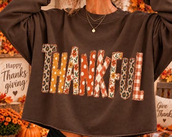 May include: A dark brown long-sleeve sweatshirt featuring the word THANKFUL across the chest. Each letter is filled with a different pattern, including leopard print, orange polka dots, floral motifs, and a red and white gingham check pattern.
