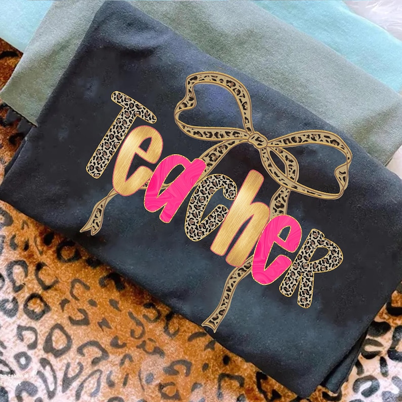May include: Black t-shirt with the word "Teacher" in gold and pink, accented with a leopard print bow. The shirt is folded, with two other shirts in shades of green visible in the background. The background has a leopard print pattern.