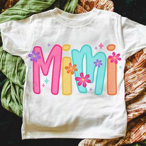 Peut inclure: Un t-shirt blanc avec le mot "Mimi" &eacute;crit en lettres color&eacute;es en forme de bulles. Les lettres sont d&eacute;cor&eacute;es de petites fleurs color&eacute;es.