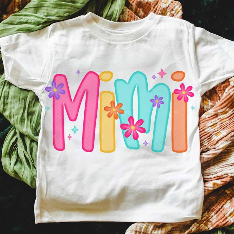 Peut inclure: Un t-shirt blanc avec le mot "Mimi" &eacute;crit en lettres color&eacute;es en forme de bulles. Les lettres sont d&eacute;cor&eacute;es de petites fleurs color&eacute;es.