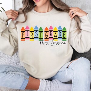 Op de afbeelding: Een cr&egrave;mekleurig sweatshirt met een rij kleurpotloden die "FIRST GRADE" spellen en de tekst "Mrs. Jessica" eronder.