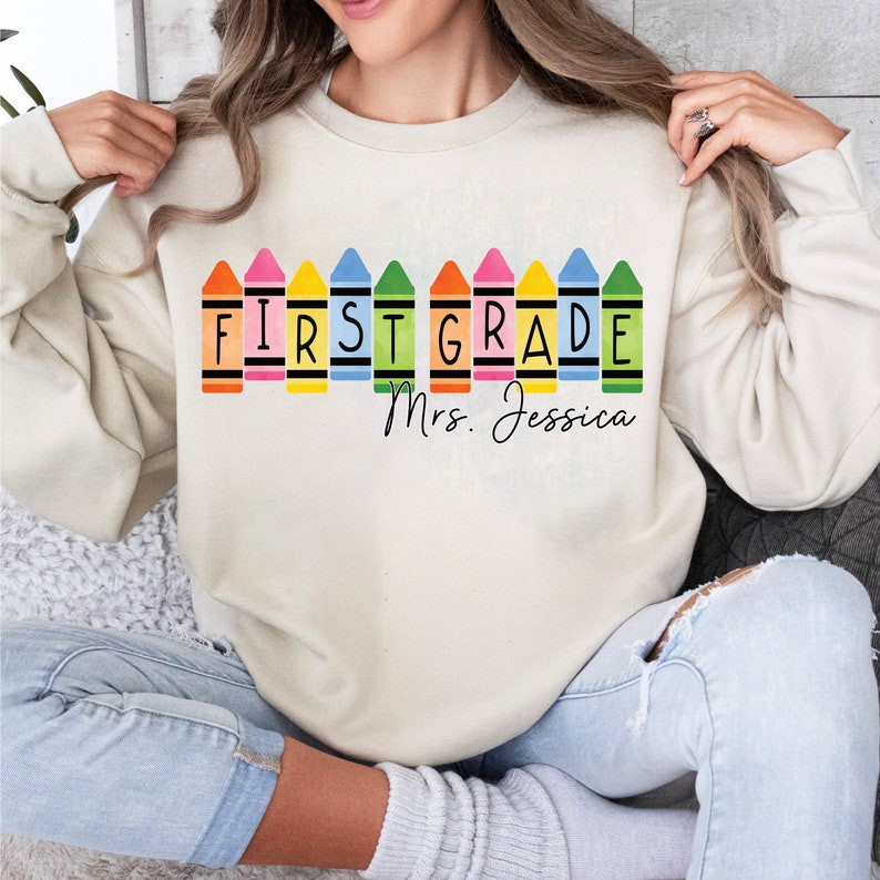 Op de afbeelding: Een cr&egrave;mekleurig sweatshirt met een rij kleurpotloden die "FIRST GRADE" spellen en de tekst "Mrs. Jessica" eronder.
