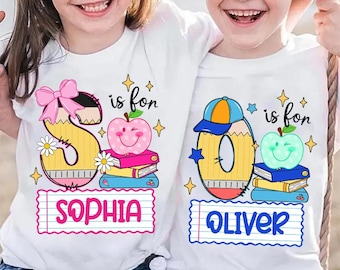 May include: Two white t-shirts featuring school-themed graphics. The left shirt displays a large letter S designed like a pencil with a pink bow, a pink apple, books, and the name SOPHIA. The right shirt shows a letter O, a blue cap, a green apple, and OLIVER.