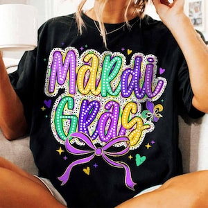 May include: Black t-shirt with a colourful "Mardi Gras" design featuring purple, green and gold glitter accents. The design includes a bow and a fleur-de-lis.
