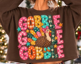 May include: A brown long-sleeve sweatshirt featuring a colorful graphic print with the text GOBBLE GOBBLE GOBBLE. The design includes a turkey illustration with a pink bow, surrounded by various patterns like polka dots and plaid.
