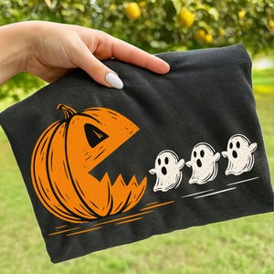 May include: Black t-shirt featuring a Halloween-themed design. An orange pumpkin with a Pac-Man-like mouth chases three white ghosts. The design is printed on a black t-shirt, perfect for the spooky season.