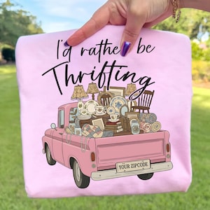 May include: Pink graphic t-shirt with the text "I'd rather be Thrifting." The design features a pink vintage pickup truck filled with lamps, dishes, and other thrifted items. The truck bed has a variety of items, including lamps, dishes, and other thrifted items.