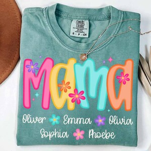 Peut inclure: Un t-shirt vert sarcelle avec le mot "Mama" en lettres color&eacute;es avec des fleurs. Les noms Oliver, Emma, Olivia, Sophia et Phoebe sont &eacute;crits sous le mot "Mama" avec des fleurs entre chaque nom.