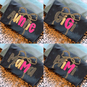 Könnte beinhalten: Vier schwarze T-Shirts mit verschiedenen Textdesigns. Jedes Shirt hat eine goldene Leopardenmuster-Schleife und einen Namen in Pink. Die Namen sind "Auntie", "Gigi", "Mama" und "Mimi". Die Shirts befinden sich auf einem Leopardenmuster-Hintergrund.