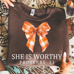 May include: A dark brown sweatshirt featuring a watercolor-style orange and white plaid bow. The text "SHE IS WORTHY PROVERBS 31" is printed in white. The sweatshirt is held up by a person.