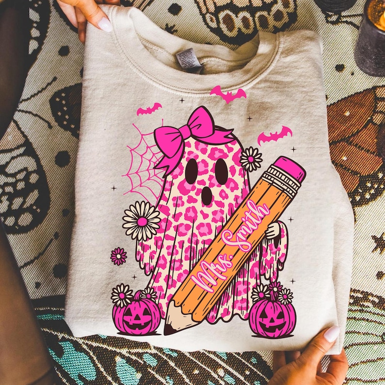 May include: A beige sweatshirt featuring a pink leopard print ghost with a bow, holding a pencil with "Mrs. Smith" on it. Pink bats, flowers, and jack-o'-lanterns complete the Halloween-themed design.
