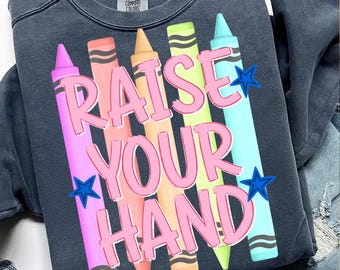 May include: A charcoal gray crewneck sweatshirt featuring a graphic print of five colorful crayons in pink, coral, orange, lime green, and light blue. The text "RAISE YOUR HAND" is printed in large pink letters over the crayons with two blue stars.