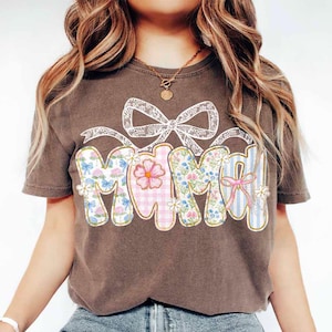 K&ouml;nnte beinhalten: Ein braunes T-Shirt mit Patchwork-Design, auf dem "Mama" in einem floralen Druck mit einer wei&szlig;en Schleife aus Spitze steht.