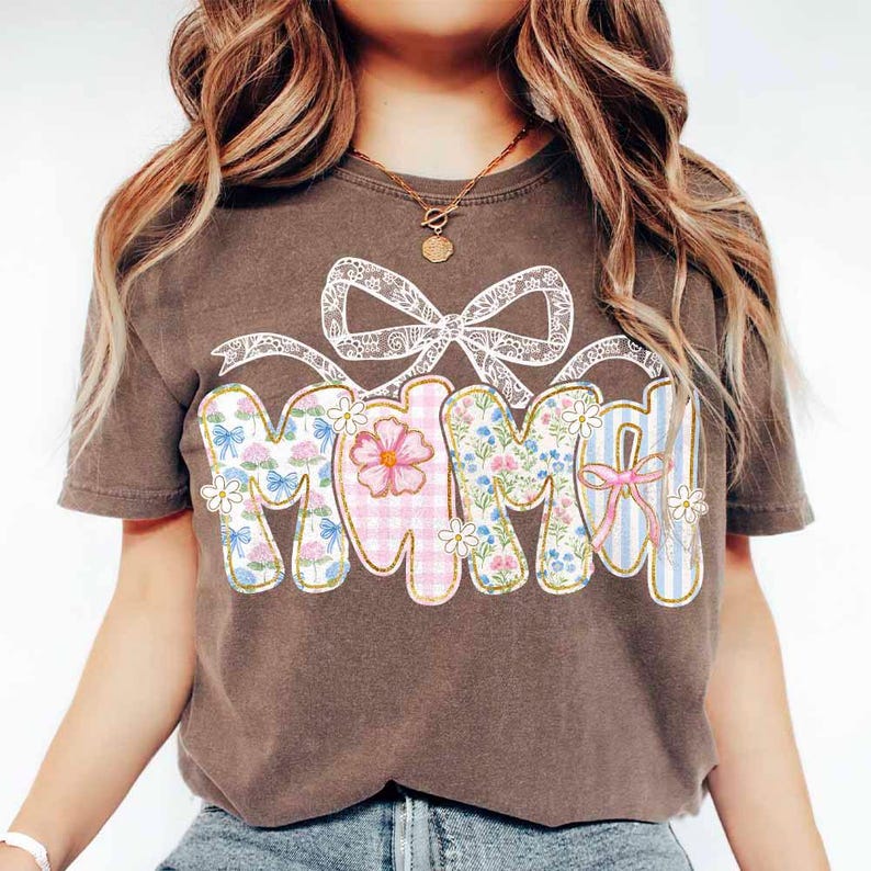 K&ouml;nnte beinhalten: Ein braunes T-Shirt mit Patchwork-Design, auf dem "Mama" in einem floralen Druck mit einer wei&szlig;en Schleife aus Spitze steht.