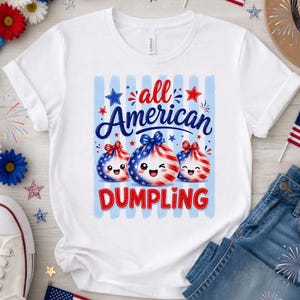 Op de afbeelding: Wit T-shirt met de tekst "all American DUMPLING" in rood en blauw. Het ontwerp toont drie dumpling illustraties met patriottische rood, wit en blauwe patronen, sterren en strikken. Het shirt wordt gecombineerd met een blauwe jeans.