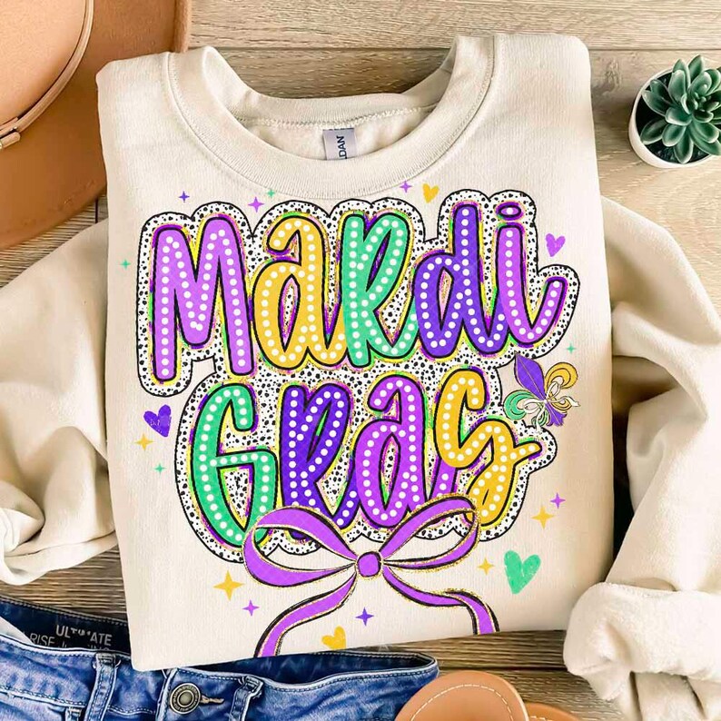 May include: A beige sweatshirt with a colourful "Mardi Gras" graphic. The graphic features purple, green and gold colours with a bow and hearts.