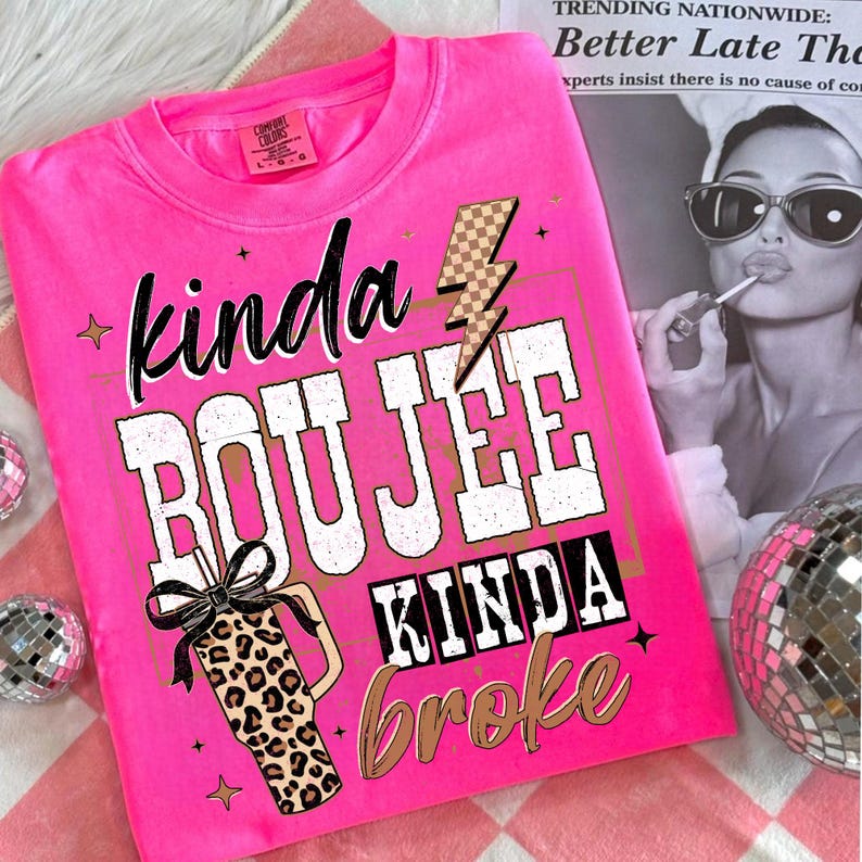 May include: A bright pink t-shirt with the words "kinda BOUJEE kinda broke" in a mix of fonts and colors. The design includes a lightning bolt, stars, and a leopard print tumbler with a bow. The shirt is a Comfort Colors brand.