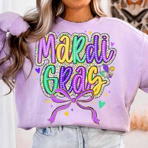 May include: A purple sweatshirt with a colourful "Mardi Gras" graphic. The graphic features a green, yellow, and purple font with white polka dots. A gold ribbon with a bow is below the text.