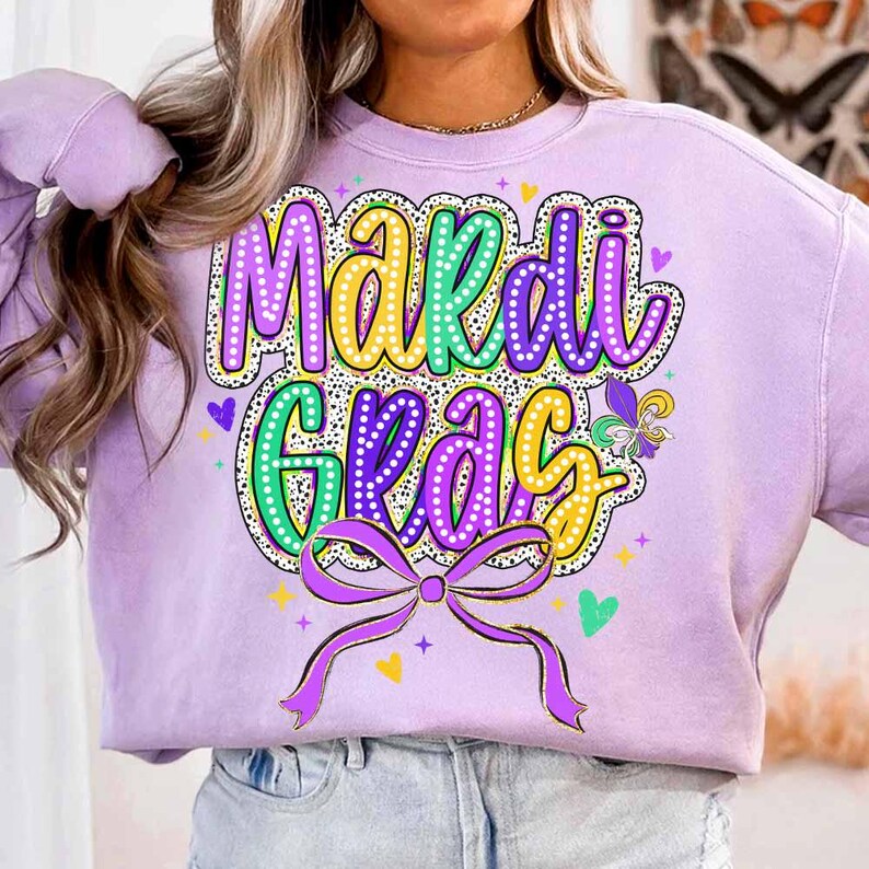 May include: A purple sweatshirt with a colourful "Mardi Gras" graphic. The graphic features a green, yellow, and purple font with white polka dots. A gold ribbon with a bow is below the text.