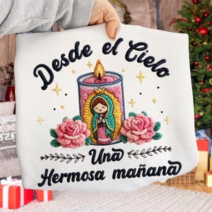 May include: White embroidered sweatshirt featuring a candle with a religious figure, pink roses, and text in Spanish. The design includes the phrases "Desde el Cielo", "Una", and "Hermosa mañana" in dark blue script.