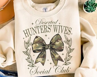 May include: A cream-colored sweatshirt with the text "Deserted Hunters' Wives Social Club" in brown lettering. A green camouflage bow with a white outline is centered on the sweatshirt.