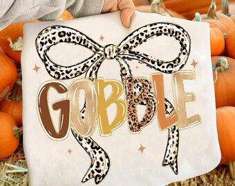 May include: A cream-colored graphic featuring the word "GOBBLE" in brown, yellow, and orange hues, with a leopard print bow. The design includes small star accents, perfect for Thanksgiving or fall celebrations.