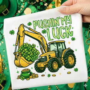 Pode incluir: Camiseta branca com um design de escavadeira amarela pegando trevos verdes. O texto "PUSHIN' MY LUCK" está em verde. Uma panela de ouro e moedas também são mostradas, com trevos e estrelas.