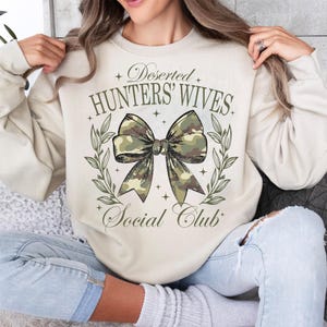 Op de afbeelding: Een beige sweatshirt met de tekst "Deserted Hunters' Wives Social Club" en een camouflage strik met groene bladeren.