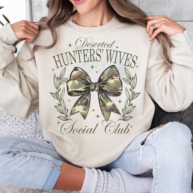 Op de afbeelding: Een beige sweatshirt met de tekst "Deserted Hunters' Wives Social Club" en een camouflage strik met groene bladeren.