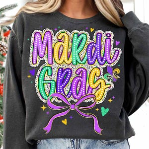 May include: A black sweatshirt with a purple, green and yellow Mardi Gras design. The design features the words "Mardi Gras" with a purple bow and gold glitter accents.